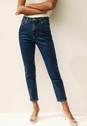 Kõrgelt taljes tumesinised denim teksad, sirge lõikega, eestaskutega ja toore äärega, kaunistatud selgete kõrgetel kontsadel kingadega.