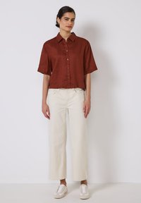 Rust-colored short-sleeve button-up shirt, relaxed fit, paired with wide-leg white trousers, and casual light shoes, on a neutral background.