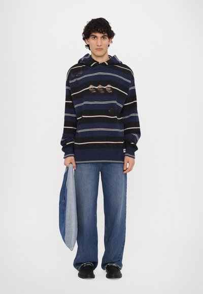 Un hoodie rayé marine en matériau tricoté avec des détails effilochés, associé à un jean en denim bleu ample et des chaussures noires, tenant un tissu gris.