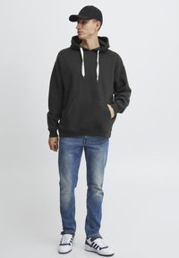 Felpa nera con coulisse bianche e tasca frontale, abbinata a jeans azzurri chiari e sneakers bianche con dettagli blu navy, su uno sfondo neutro.