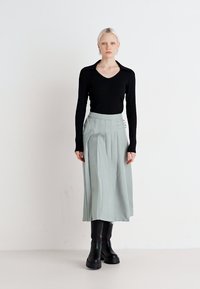 Must sabi mustega mustega long-sleeve top mustega paired with light green pleated skirt featuring buttons and a high waist. Model wears chunky black knee-high boots.