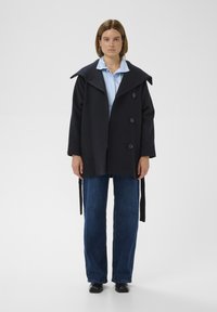 Kabát z námorníckej vlny so širokým golierom, dvojradové gombíky, bočné viazanie, nosený cez svetlomodrú košeľu a tmavé denimové nohavice. Čierne topánky.