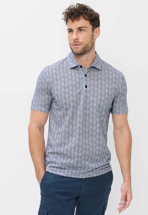 Mann mit lockigem Haar, der ein graues, gemustertes Polo mit kurzen Ärmeln und dunkelblaue Hose trägt, steht mit einer Hand in der Tasche.