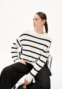 Gestreifter Pullover in Schwarz und Weiß mit weitem Rundhalsausschnitt, strukturiertem Strick und langen Ärmeln. Kombiniert mit weiten, schwarzen Hosen.