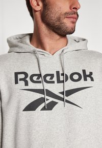 Vyras su šviesiai pilku gobtuvu, ant kurio juodas Reebok logotipas ir pintinės, stovintis šonu prieš paprastą foną.