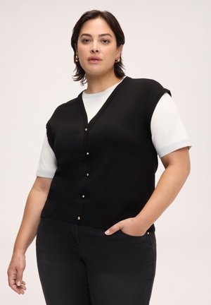 Frau trägt eine schwarze ärmellose, geknöpfte Weste über einem weißen Kurzarmhemd, schwarze Hose und goldene Kreolen-Ohrringe, steht mit der Hand in der Tasche.
