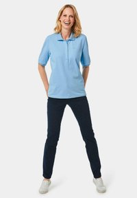 Helles blaues Polohemd mit kurzen Ärmeln und einer Knopfleiste, kombiniert mit dunkelblauen Jeans und grauen Slippern.