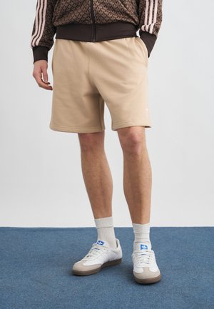 Άνδρας με μπεζ σορτς, λευκά αθλητικά παπούτσια Adidas, λευκές κάλτσες και καφέ μπουφάν με ροζ ρίγες, στέκεται πάνω σε μπλε μοκέτα.