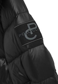 Rękaw czarnej puchowej kurtki z teksturowaną tkaniną, pikowanym wzorem oraz czarną naszywką z wypukłym szarym napisem "POLO CLUB".