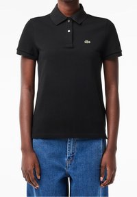 Personne portant un polo noir avec un petit logo crocodile vert sur la poitrine gauche et un jean bleu en denim.