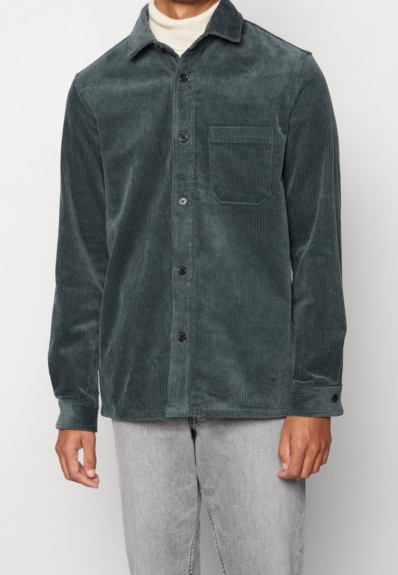 Uomo che indossa una camicia button-up in velluto a coste verde scuro sopra un maglione bianco a collo alto con jeans grigio chiaro.