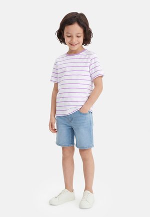 Niño con cabello rizado y oscuro que lleva una camiseta de rayas blancas y lavanda, pantalones cortos de mezclilla y zapatillas blancas, sonriendo con una mano en el bolsillo.