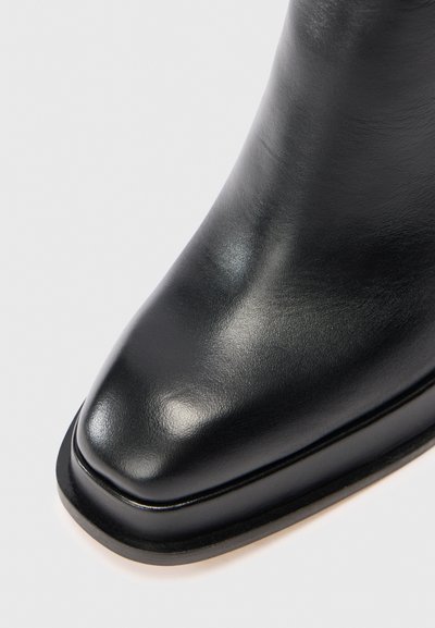 Botín negro de cuero con una textura suave y brillante, diseño de puntera cuadrada y costuras mínimas. Presenta una suela delgada, completamente negra.