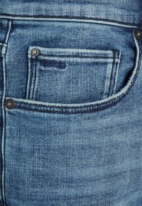 Blå denimjeans med en bakficka, urtvättad textur och kontrasterande sömmar. Silverknapprande detalj synlig på fickan.