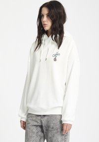Haut en coton blanc avec une poche kangourou à l'avant, capuche à cordons, et un motif brodé vert et bordeaux sur la poitrine gauche.