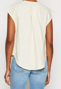 Top corto de color beige claro con dobladillo redondeado y pliegues en la parte trasera. La tela parece suave y texturizada, combinado con unos jeans de color azul claro.