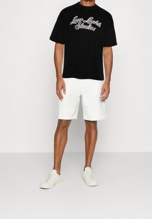 Man wearing black "Low Lights Studios" t-shirt, white knee-length shorts, and white sneakers, standing against a plain light background.
