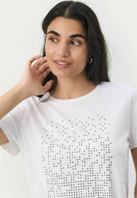 T-shirt bianca in cotone con un motivo a pois di piccoli chiodi neri e argento nella parte anteriore, con maniche corte e scollatura a girocollo.