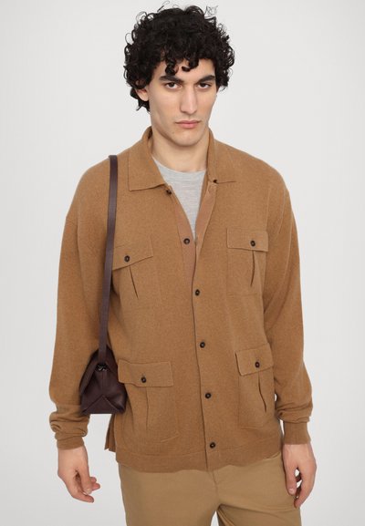 Cárdigan marrón de botones hecho de material suave, con dos bolsillos frontales, usado sobre una camisa de color gris claro; combinado con un bolso pequeño marrón.