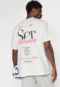 Biała bawełniana koszulka z krótkim rękawem z dużą czarno-czerwoną grafiką z napisem "Ser Benfiquista" oraz emblematem na plecach.