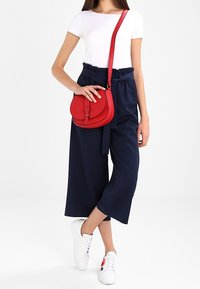Kvinde iført hvid t-shirt, navy papirsækliv bukser, hvide sneakers med blomstermotiv og bærende en rød crossbody taske.
