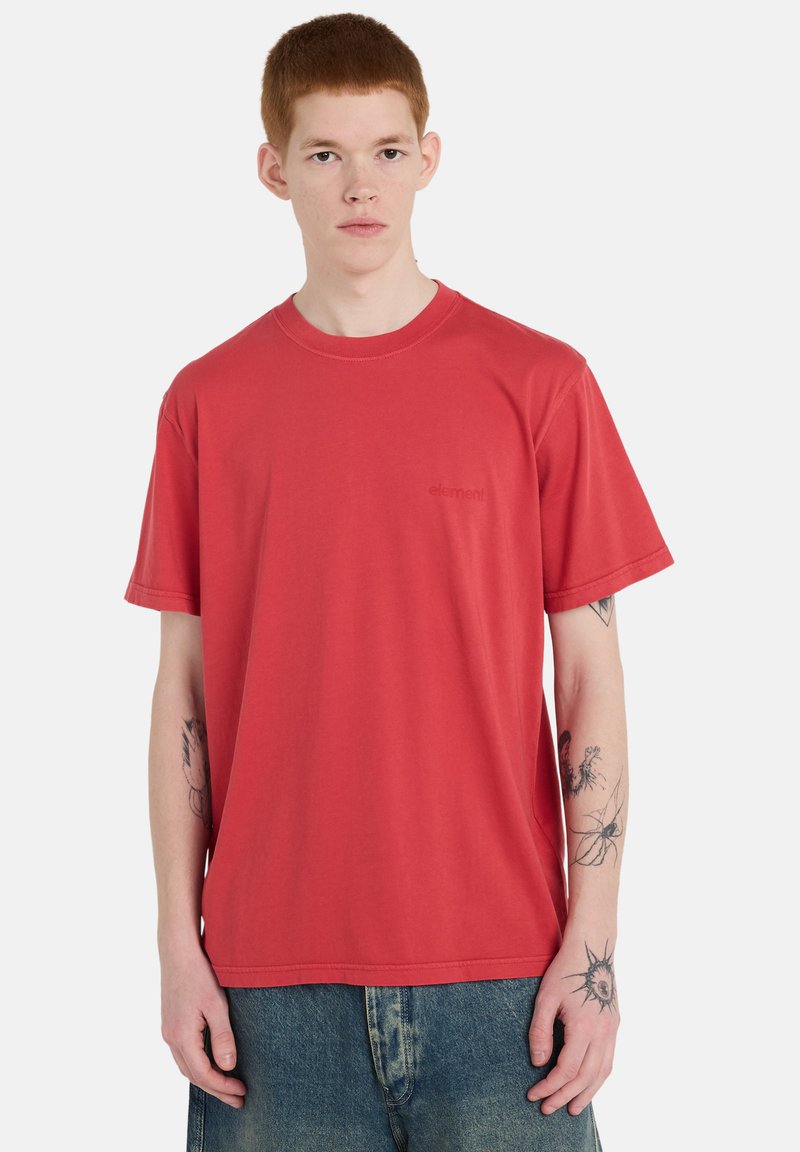 T-shirt en coton rouge à col rond et manches courtes, avec un petit logo sur la poitrine, porté avec un pantalon en denim bleu.