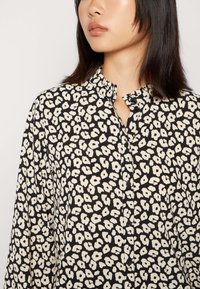 Camisa preta com um padrão floral em bege. O design apresenta um colarinho alto, fecho com botões à frente e um corte descontraído. O tecido parece leve e texturizado.