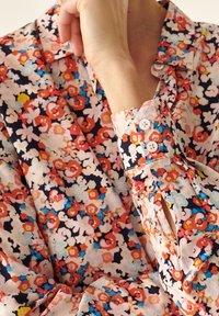 Närbild av en person som bär en blommig blus med långa ärmar i röd, rosa, blå och gul färg, som vilar sin hand nära ansiktet.