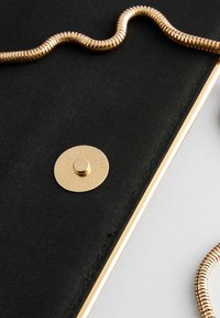 Clutch de tela negra con cierre de botón dorado y cadena dorada. Superficie texturizada con bordes cosidos, resaltando el contraste de materiales.