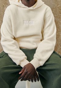 Cremefarbener Fleece-Hoodie mit strukturierter Oberfläche und einem auf der Brust aufgenähten Logo, kombiniert mit lockeren grünen Hosen.