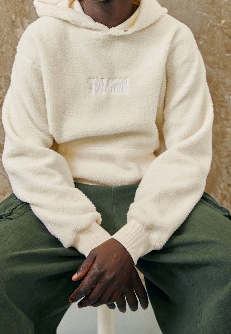 Cremefarbener Fleece-Hoodie mit strukturierter Oberfläche und einem auf der Brust aufgenähten Logo, kombiniert mit lockeren grünen Hosen.