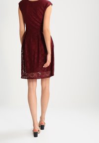 Vestido de encaje burdeos con un corsé ajustado, mangas cortas y una falda acampanada. Presenta un patrón festoneado y cae por encima de la rodilla. Tiras transparentes visibles.