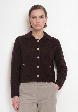 Kvinde iført en mørkebrun tekstureret jakke med guldknapper og lyse beige kordfløjlsbukser, stående med den ene hånd i lommen mod hvid baggrund.