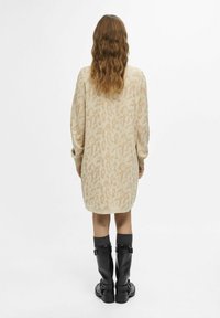 Beige vzorované svetr šaty s dlouhými rukávy, které mají texturovaný design. Nosené s černými vysokými kozačkami a šedými ponožkami.
