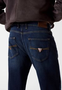 Jeans in denim blu scuro con cuciture gialle, dotati di due tasche posteriori e una patch a forma triangolare. Texture liscia, taglio a gamba dritta.