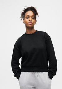 Νεαρό άτομο με σγουρά μαλλιά, φορώντας μαύρο φούτερ και ανοιχτόγριες παντελόνες με κορδόνι, στέκεται με τα χέρια στις τσέπες, ουδέτερο φόντο.