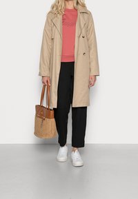 Žena v béžovém trenčkotě, korálovém svetru, černých kalhotách, bílých teniskách, držící hnědou tkanou kabelku na jednobarevném pozadí.