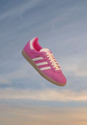 Zapatilla Adidas Samba rosa con rayas blancas y suela de goma flotando sobre un fondo de cielo suave azul y naranja.