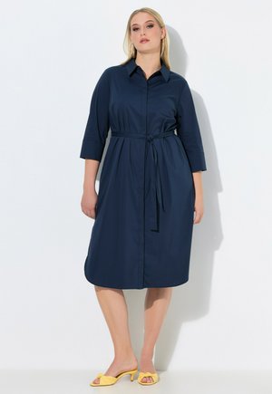 Kvinde stående iført en marineblå, knælængde kjole med bælte, 3/4 ærmer og gule åbne tå-højhælede sandaler mod en hvid baggrund.