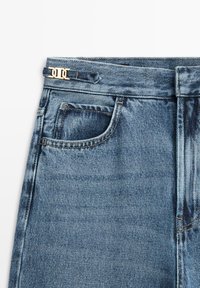 Hellblaue Jeans aus Denim mit hoher Taille, versehen mit einem goldfarbenen Logo-Beschlag am Gürtel und einer Vordertasche mit kontrastierenden Nähten.