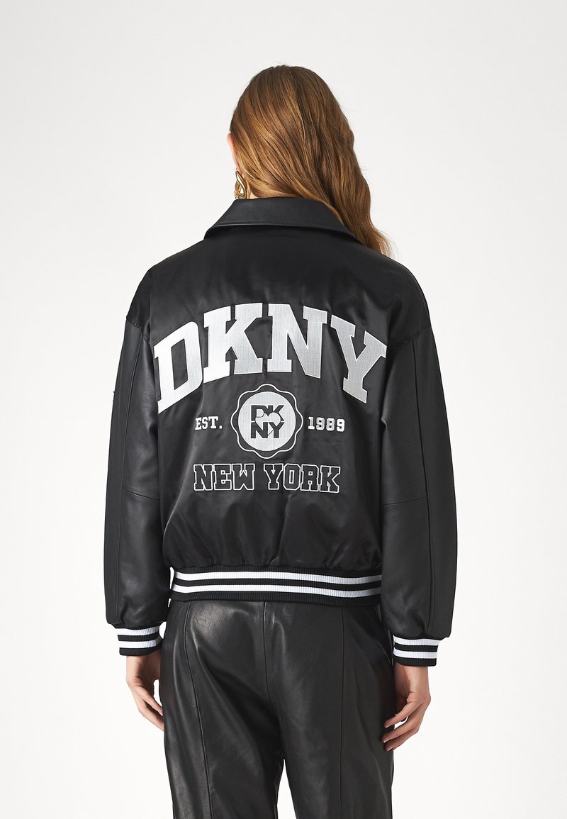 Blouson aviateur noir à finition satinée, avec inscriptions blanches DKNY et logo au dos, poignets et ourlet côtelés rayés.