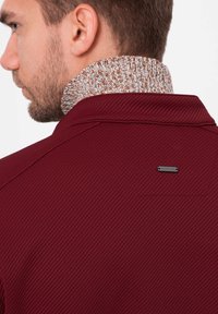 Burgunderfärgad polo tröja med texturerad tyg, med en hög stickad krage i ett brunt och gräddvitt mönster och en liten svart varumärkesetikett på baksidan.