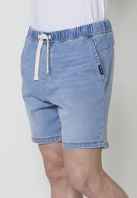 Blå denimshorts med elastisk midja och vit snodd. Inkluderar sidofickor och kontrasterande sömmar. Mjuk textur, avslappnad passform.