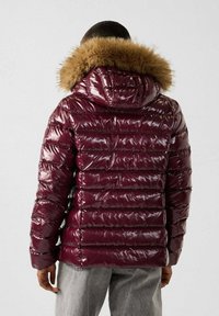 Burgunderfarbene Puffjacke mit glänzender Oberfläche, horizontaler Steppung und Kunstfellbesatz an der Kapuze. Lange Ärmel und viel Polsterung für Wärme.