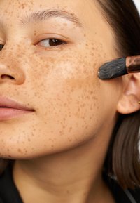 Close-up of a face with prominent freckles, applying product with a brush. Skin is smooth and has a warm tone. Hair is dark, straight, and medium length.