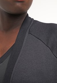 Detailní záběr dolní části obličeje a hrudníku osoby, která má na sobě tmavě šedou texturovanou bundu přes černé tričko.