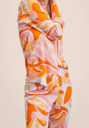 Femme portant un ensemble coordonné composé d'un chemisier à manches longues et d'un pantalon à motifs floraux audacieux en orange, rose et blanc sur un fond uni.