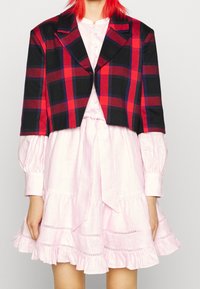 Svart och röd rutig kort jacka med notched lapels över en ljusrosa klänning med volangkant och knytning i midjan. Texturerad tyg synlig.