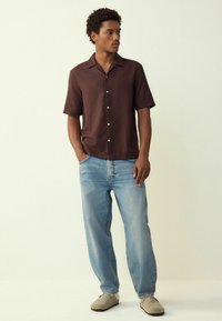 Short-sleeve brown shirt with textured pattern, button-up front. Paired with light blue wide-leg jeans and beige slip-on shoes.
