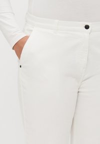 Hvite bukser laget av glatt stoff med en tettsittende design, som har lommer på siden og en svart knapplukking i midjen.